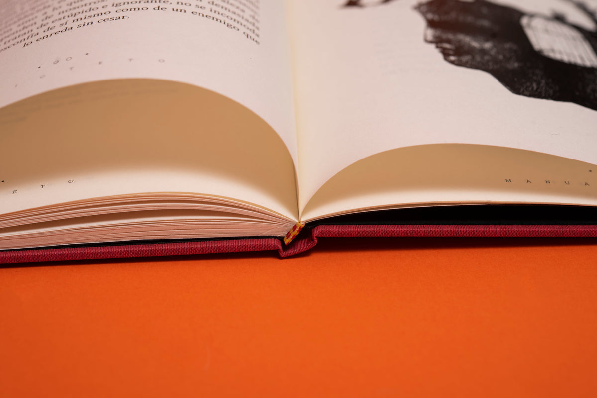 Detalle del lomo cosido a mano y de la tapa en tela vinotinto, con el libro abierto sobre fondo naranja